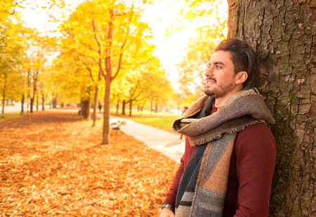 Handsome man leaning against a tree in a park in autumn while smiling
