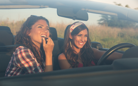 Two young beautiful girls driving and putting on lipstick in a convertible car photographed from outsideの写真素材
