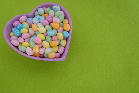 Jelly beans in a purple heart shaped bowl on a green fabric backgroundの写真素材