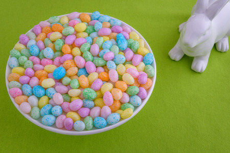 Jelly beans in a on a white cake plate on a green fabric background, with a white bunny looking onの写真素材