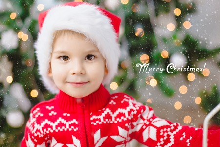 Little boy in santa claus hat looks at the camera and smile. Bokeh background. Copy-space. Text contains good wishes. Christmas. Holidayの写真素材