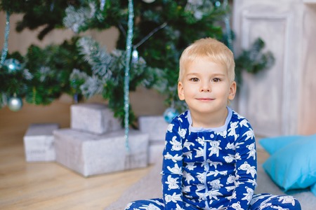 Little boy in pajamas with bears sit on the floor and smile. Hold his legs. Near christmas tree and...