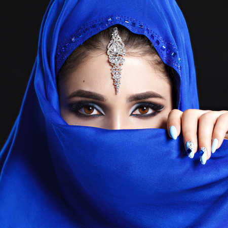 Gorgeous Young East Woman face portrait in hijab. Beauty Model Girl with bright eyebrows, perfect make-up, touching her face. Traditional. Isolated on black background. Smokey.Jewelry on her face. Studioの写真素材