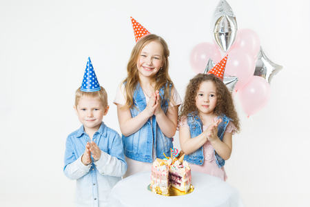 group of kids in festive caps clap their hands near birthday cake, balloons on background. Studioの写真素材