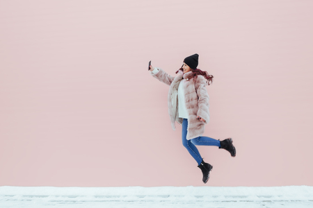 Fashion portrait of pretty smiling and jumping woman in mink coat with smartphone against the colorful pink wall. Copyspace. Flying colored hairsの写真素材