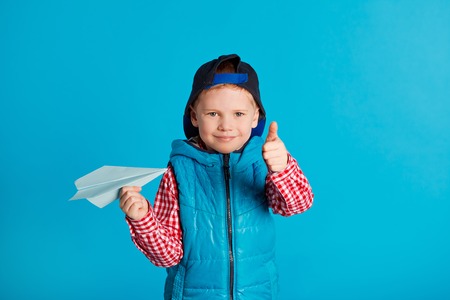 Concept travel. Fly. Little funny boy in cap playing with blue paper plane on blue backgroundの写真素材