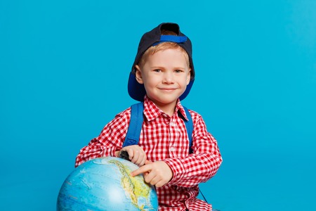 portrait of little funny boy with backpack and cap. Showing place on the globe, map. Concept travel, education. On the blue backgroundの写真素材