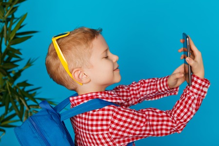 Little hipster boy with backpack and sunglasses holding telephone and making selfie. Concept travel, education, technology. Isolated on the blue backgroundの写真素材