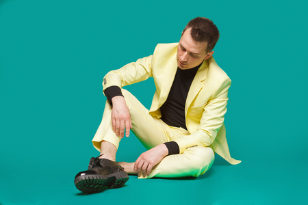 Attractive young man in suit sitting on the floor. Stylish outlook, successful businessman, happy, expressing true positive emotions, funny. Place for text. Studioの写真素材