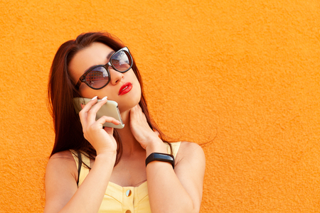 Fashion portrait of pretty smiling woman in sunglasses talking on the smartphone against the orange wall.fitness bracelet on the hand. Red lipsの写真素材