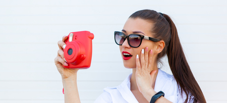 Fashion portrait of pretty smiling woman in sunglasses making photo by the pink camera against the grey wall. fitness bracelet on the handの写真素材