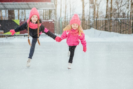 Two Little smiling girls skating on ice in pink wear and hand made scarfs. Outdoor. Winter. Lifestyle and sportの写真素材