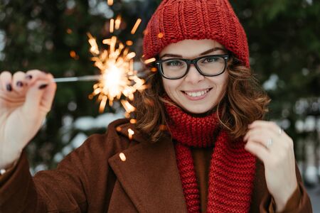Young beautiful woman in knitted red hat and scarf standing in the park with bengal light, sparkler. Concept celebration, christmas. Winterの写真素材