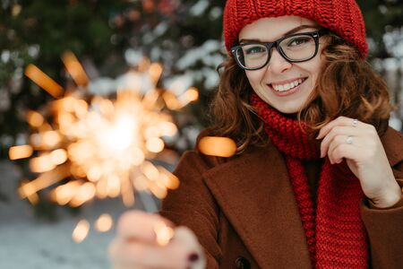 Young beautiful woman in knitted red hat and scarf standing in the park with bengal light, sparkler. Concept celebration, christmas. Winterの写真素材