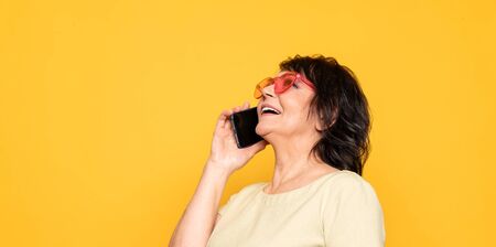 Beautiful Senior woman in pink neon sunglasses talking by the smartphone over yellow background. Professional make-upの写真素材