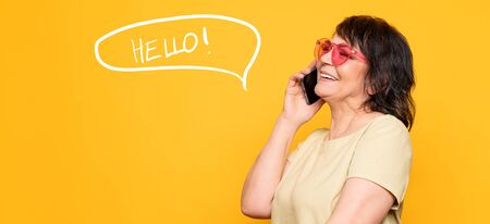 Beautiful Senior woman in pink neon sunglasses talking by the smartphone over yellow background. Professional make-upの写真素材