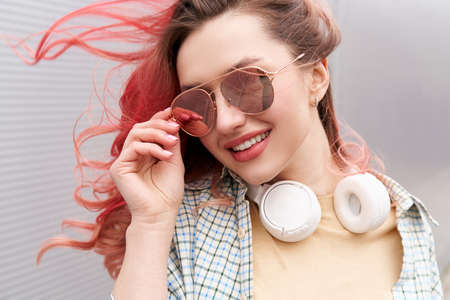 Young hipster woman with flying coloured hairs smiling in sunglasses, white headphones on her neck