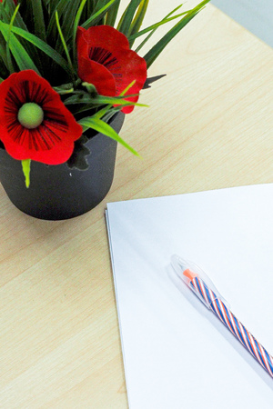 pen lay on blank white paper beside black pot of fake red flowers and green leaves put on wooden tableの写真素材