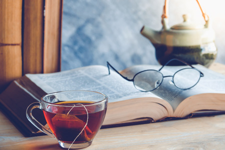 Open old book  and reading glasses above the text with tea cup and clock on tableの写真素材