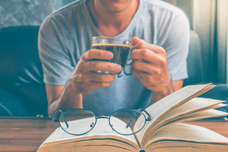 Young man is sitting on a sofa and reading a book while holding a cup of coffee