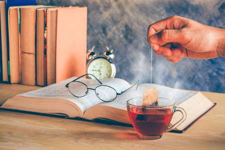 Hand of man dripping tea bag in a tea cup on table with booksの写真素材