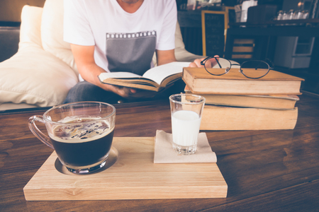 Young man is sitting on a sofa and reading a book and  cup of coffee on table in cafeの写真素材