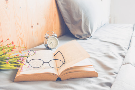 Open old book with glasses  and alarm clock on bed at morning timeの写真素材