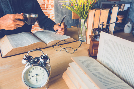 A man holding a pen and holding cup of coffee with old book on tableの写真素材