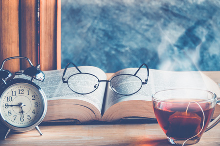 Open old book  and reading glasses above the text with tea cup and clock on tableの写真素材