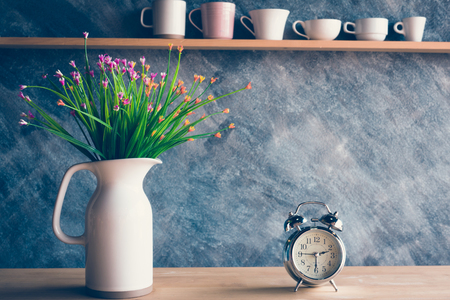 Vintage alarm clock with flower in jug on table and various cups backgroundの写真素材