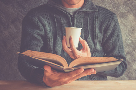 Young man is sitting on a table and reading a book while holding a paper cupの写真素材