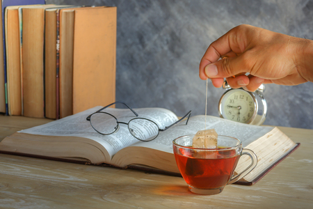Hand of man dripping tea bag in a tea cup on table with booksの写真素材