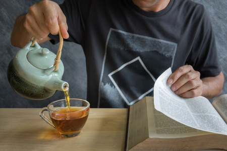 A man reading text book with pouring tea form teapot  into the cup on wood tableの写真素材