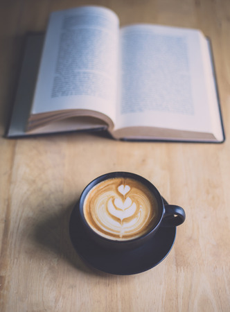 Latte art coffee in black cup with open book on wood table in coffee shopの写真素材