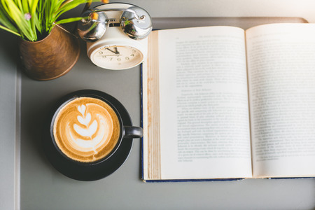 Top view of a cup of Latte coffee with open book and alarm clock with flower on table in coffee shopの写真素材