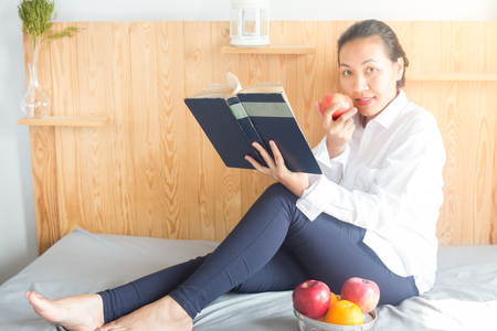 Asian young woman reading book and eating red apple on bedの写真素材