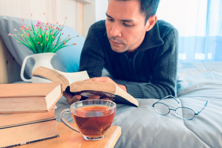 A man reading book on bed with cup of tea and stack book on tableの写真素材