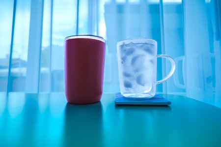 Food Series : A red thermal mug and a clear glass filled with cold water on a white table in a cozy atmosphere. White table in a clean room with natural light. A place to sip coffee with a relaxing atmosphere.の写真素材