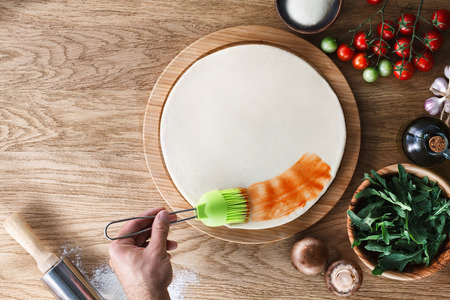 Pizza cooking process: spreading tomato sauce on pizza base with a silicone pastry brush. Wooden background, top view.の写真素材