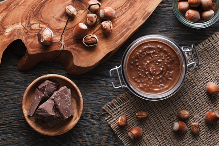 Glass jar with some hazelnut spread, a wooden bowl with milk chocolate, cutting board and nuts on a dark wooden background.の写真素材