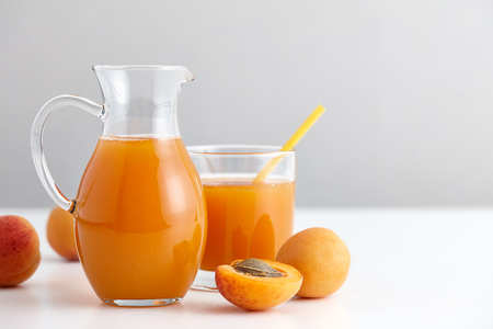 A glass jug with fresh apricot juice and ripe fruits on a white table, gray background.の写真素材
