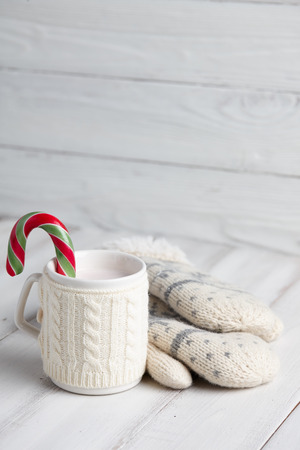 Knitted winter cup of hot chocolate with a candy cane and mittens on white wooden backgroundの写真素材