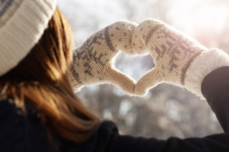 Young woman in winter mittens holding hands in a heart shape against the sunlight, feelings conceptの写真素材
