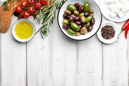 Fresh olives, feta cheese, cherry tomatoes, chili, rosemary, garlic, peppercorns and olive oil on white wooden backgroundの写真素材