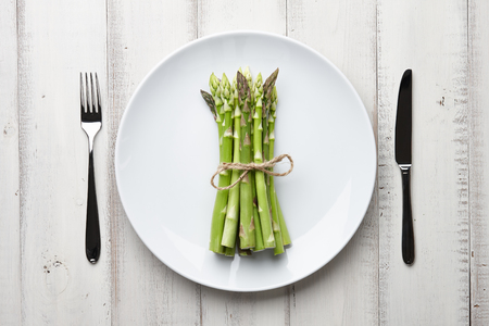 Dinner plate with bunch of fresh asparagus, fork and knife on white wooden background, top viewの写真素材