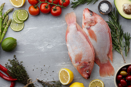 Raw red tilapia fish cooking with herbs, spices, green asparagus, tomatoes, lemon, lime and chili pepper on gray stone background, copyspaceの写真素材