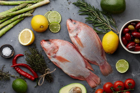 Raw red tilapia fish cooking with herbs, spices, green asparagus, avocado, lemon, lime, olives and chili pepper on gray stone background, top viewの写真素材