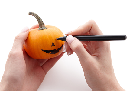 Womans hands drawing scary face on little pumpkin with a black marker pen isolated on white background for Halloween. Holiday decoration concept.の写真素材