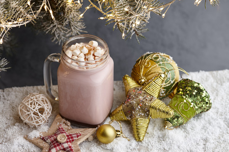 Glass winter cup of hot chocolate drink with marshmallow and Christmas toys on snowy surface under a fir tree with lightsの写真素材