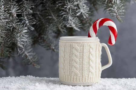Knitted winter cup of hot chocolate with candy cane on snowy table under christmas treeの写真素材